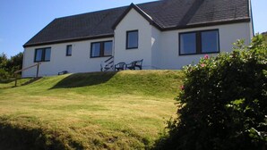 A two-story house with a sloped roof, a front porch with a bench, and a well-maintained lawn. - Steading Holidays - Eilean Feoir (Acharacle)