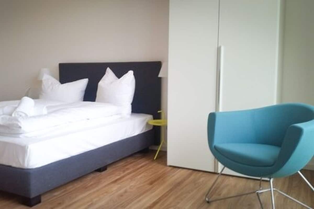Uma cama bem arrumada com lençóis brancos, uma cabeceira escura e uma cadeira azul em um quarto moderno com piso de madeira.