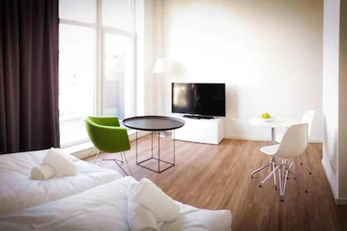 Quarto de hotel com cama, uma cadeira verde, uma mesinha redonda, televisão e escrivaninha branca.