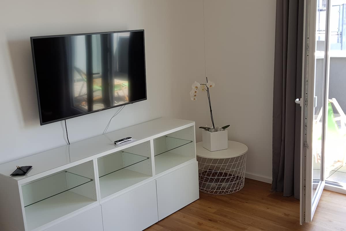 Uma sala de estar moderna com uma televisão de tela plana fixada em um móvel branco, uma pequena orquídea em vaso sobre uma mesa redonda, e uma porta de vidro deslizante que dá para uma área externa.