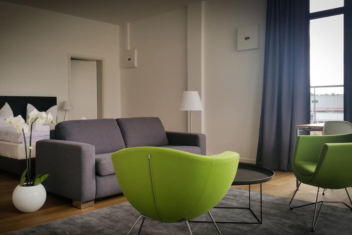 Quarto de hotel moderno com sofá cinzento, poltronas verdes, cama com almofadas brancas e flores, uma mesinha e vista pela janela.
