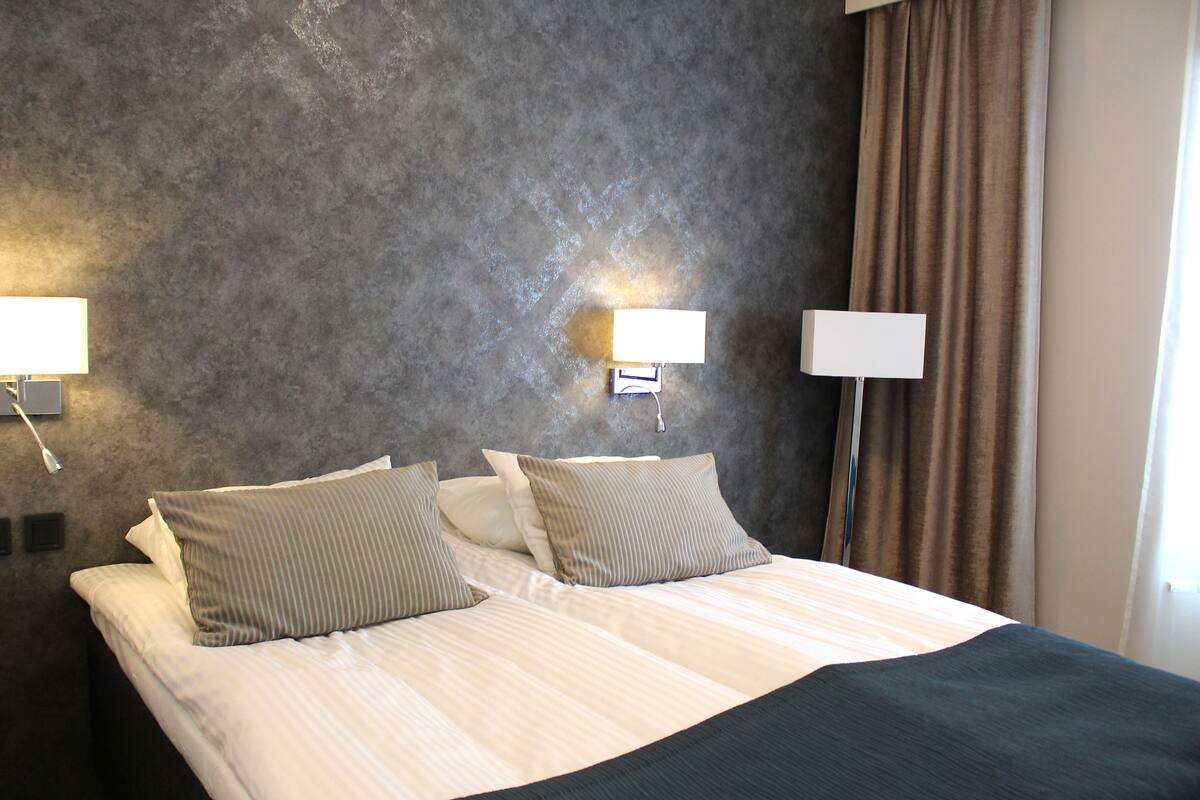 Habitación de hotel con una cama grande, dos lámparas de noche, cabecero oscuro y una ventana con cortinas.
