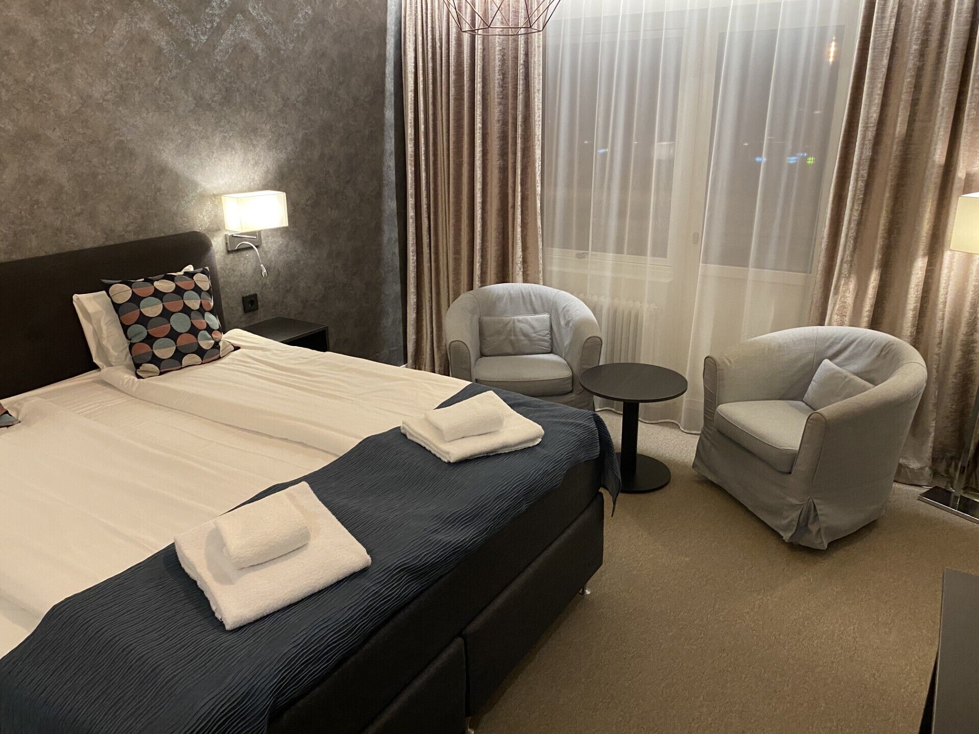 A hotel room with a large bed, two armchairs, a small round table, and a nightstand with a lamp.