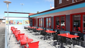 An outdoor cafe with red and black tables and chairs, a blue and red building facade, and a clear sky. - Hotell Rättvik (Rattvik)