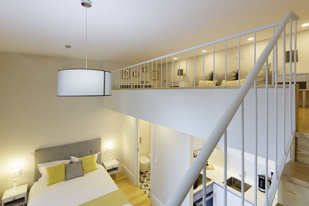A bedroom with a bed, bedside tables, a nightstand, a hanging light fixture, a staircase, and a bathroom visible through an open door.