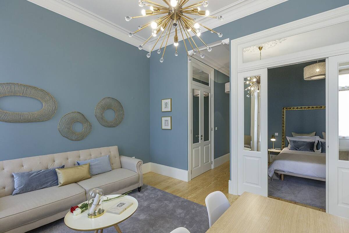 A living room with a beige sofa, a round coffee table, and a chandelier. There is a door leading to a bedroom with a bed and a mirror.