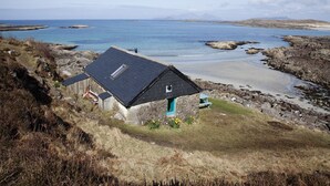 Ein kleines Steinhaus mit dunklem Dach, gelegen auf einem grasbewachsen Hügel mit Blick auf einen Strand und das Meer.
