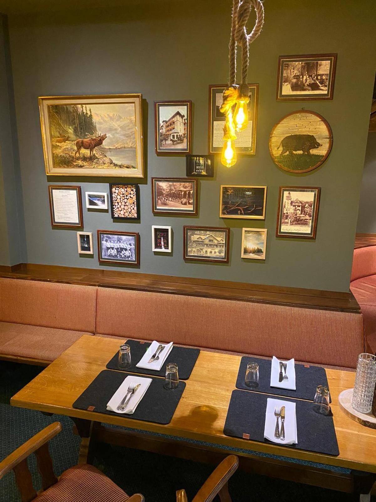 Un comedor con una mesa de madera dispuesta para dos personas, un banco y una pared decorada con cuadros y una lámpara colgante.