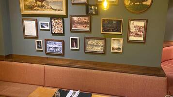 Un coin repas avec une table en bois dressée pour deux personnes, un banc et un mur décoré de tableaux encadrés et d’un luminaire suspendu.