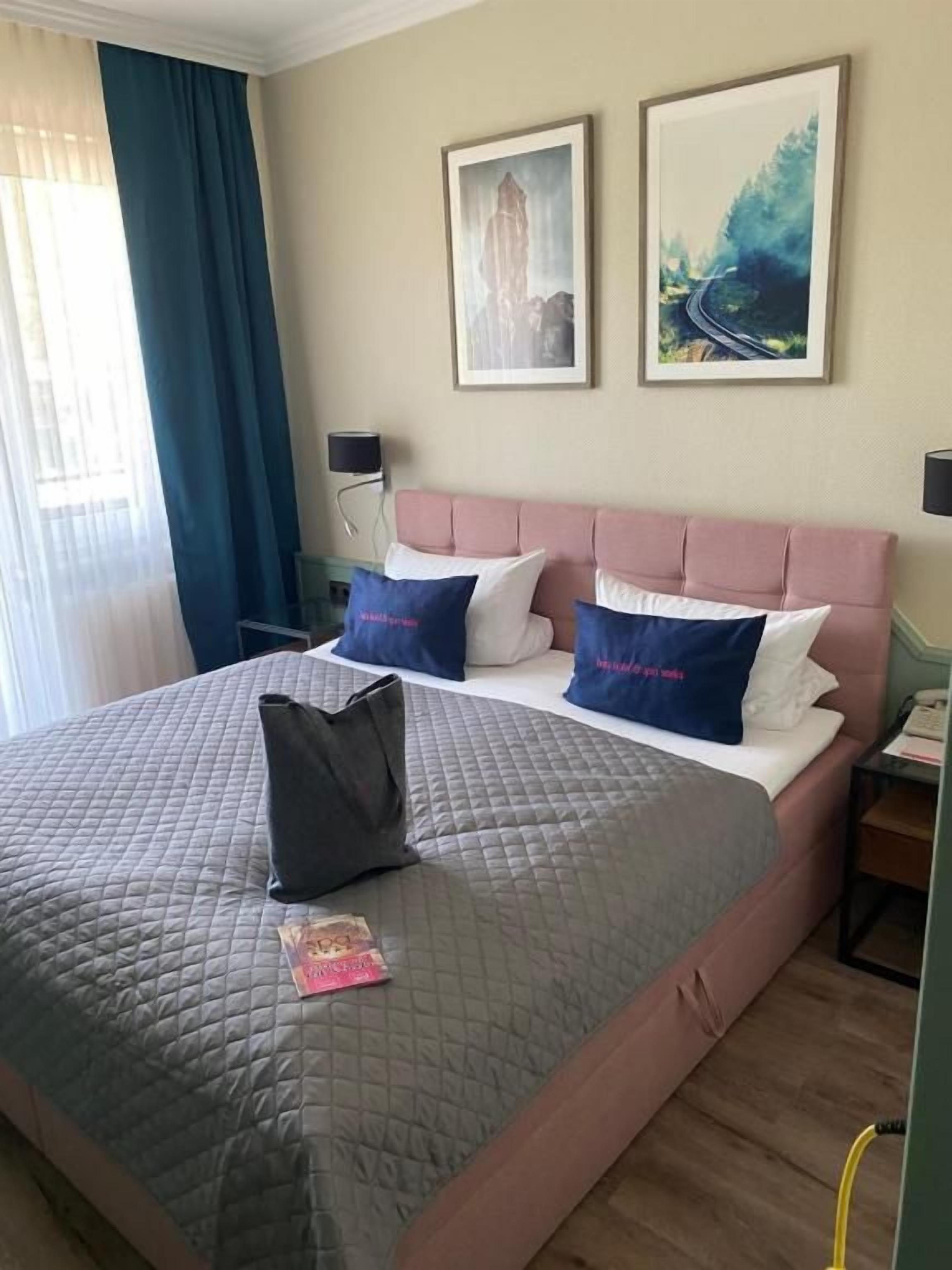 A hotel room with a bed, two pillows, a bedside table, a lamp, and a window with curtains.