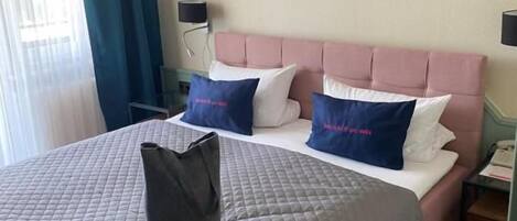 A hotel room with a bed, two pillows, a bedside table, a lamp, and a window with curtains.