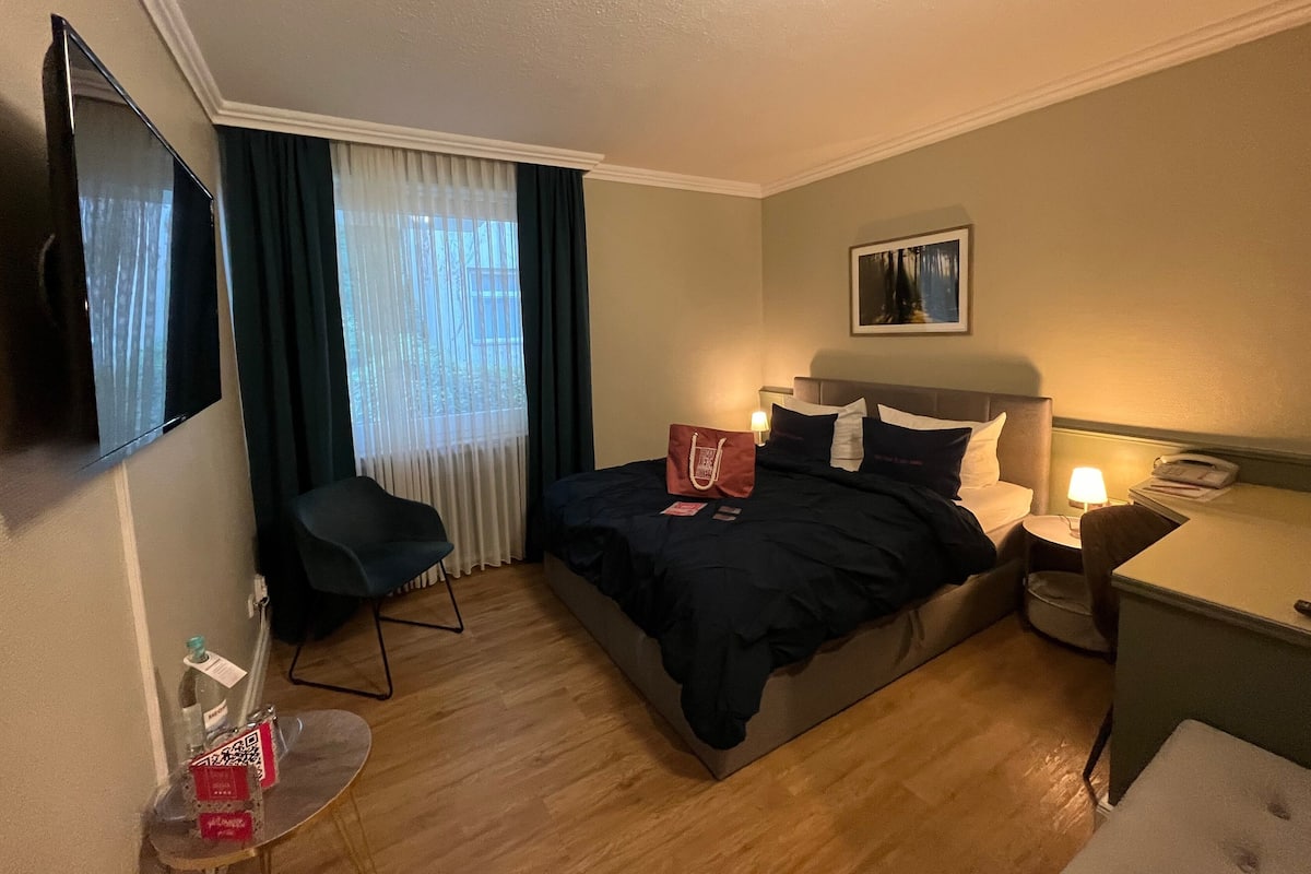 A hotel room with a bed, a desk, a chair, a TV, and a window with curtains.