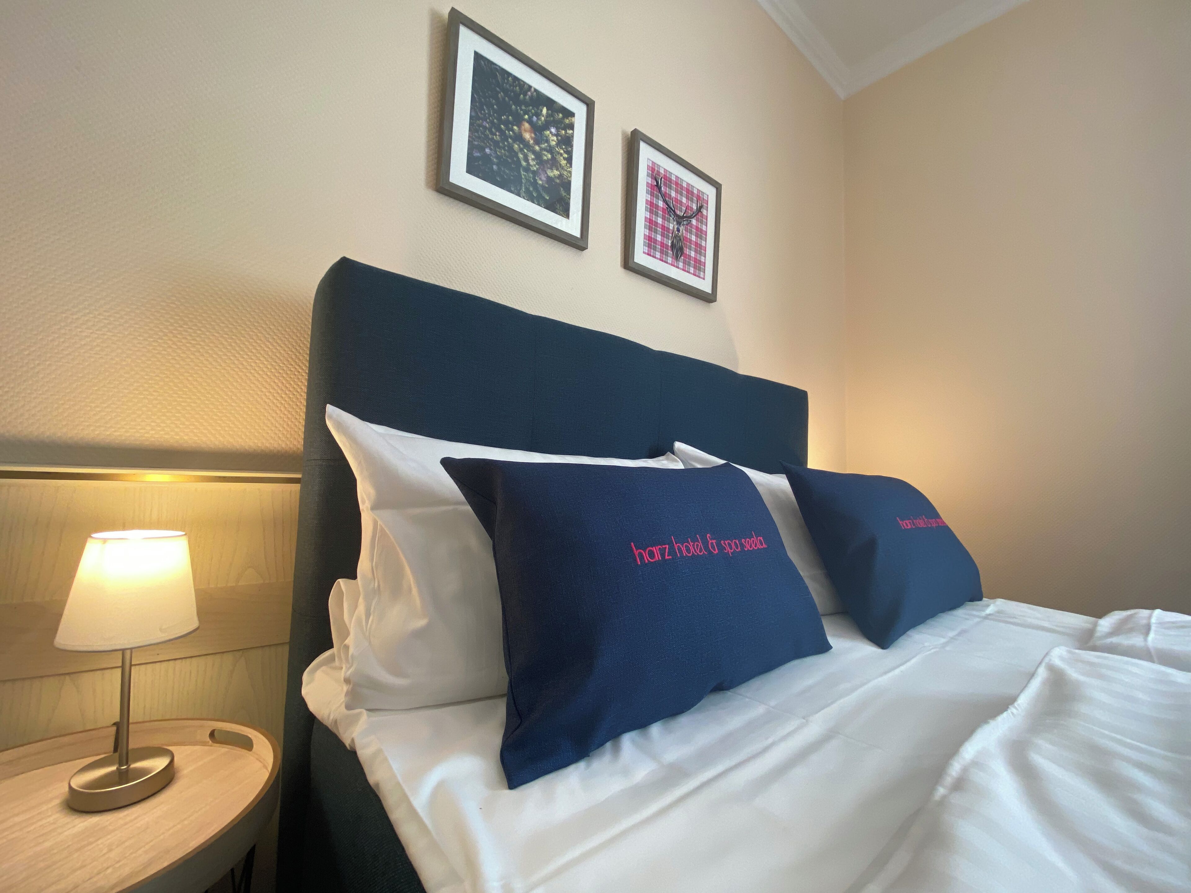 A neatly made bed with white and navy blue bedding, two framed pictures on the wall, and a bedside lamp on a wooden nightstand.