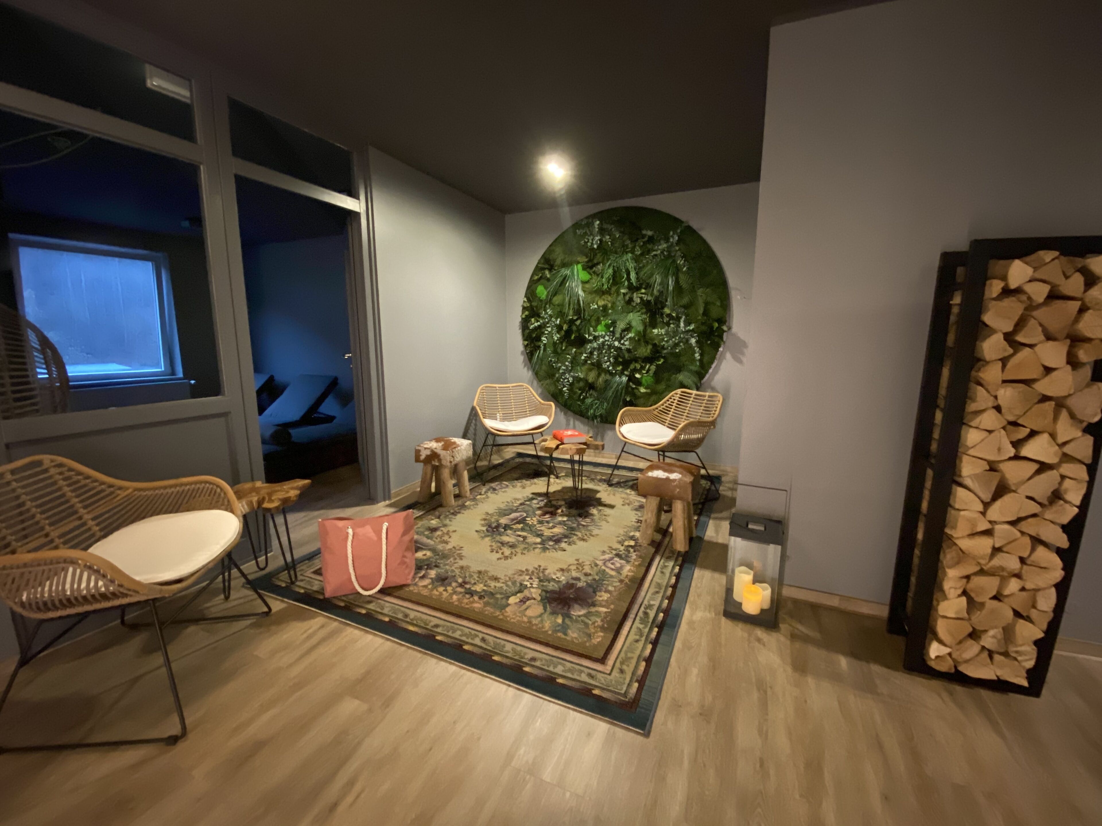 A modern living room with a large round green wall decoration, wicker chairs, a patterned rug, and a stack of firewood.