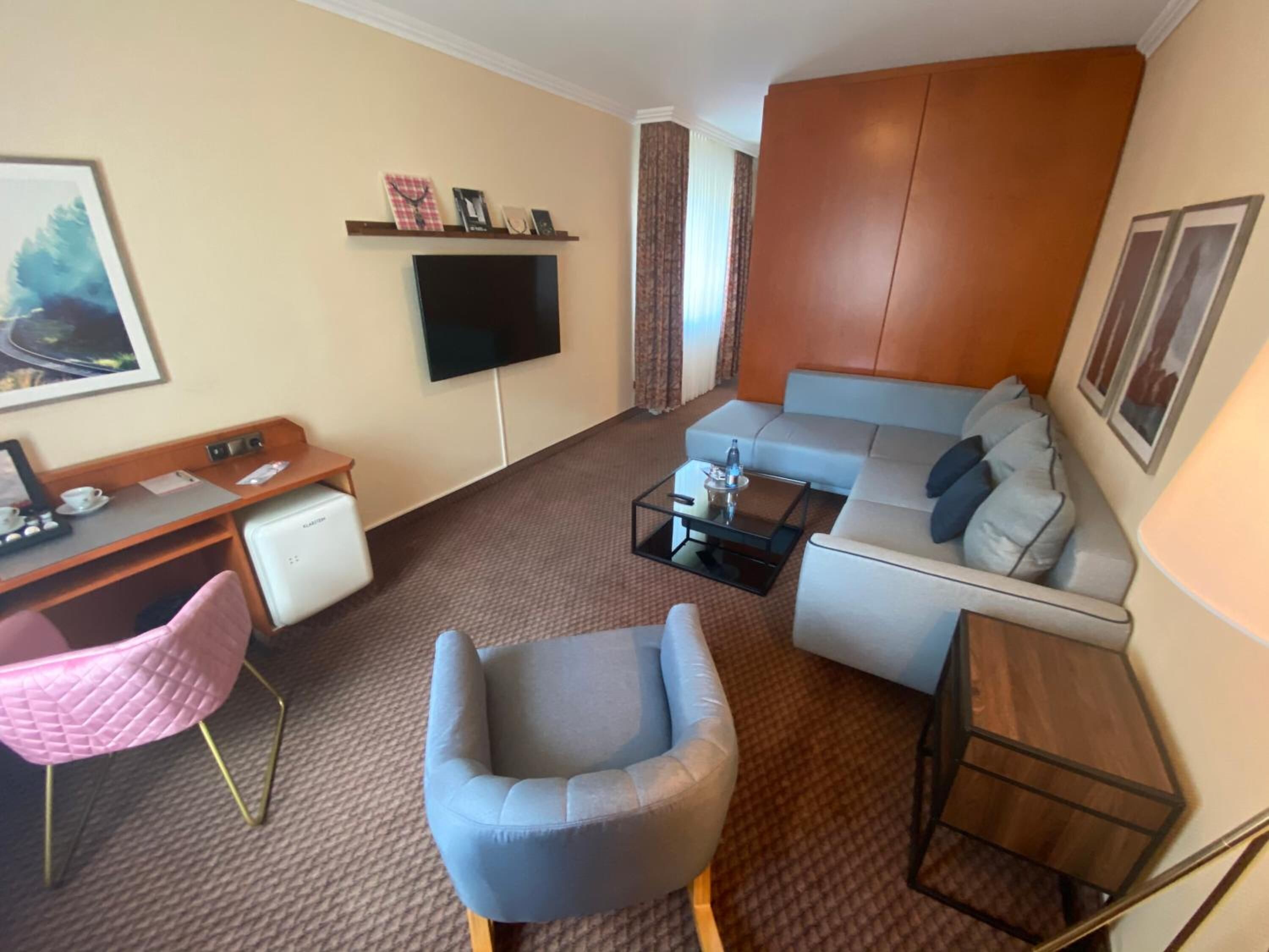 Een gezellige, compacte woonkamer met een bank, fauteuil en een klein bijzettafeltje. Een flatscreen televisie is aan de muur bevestigd, en er staat een bureau met een stoel.