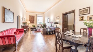 Amplia sala de estar con un sofá rojo, una mesa de comedor de madera y sillas. Hay una chimenea, un ventanal grande y varios objetos decorativos en las paredes.
