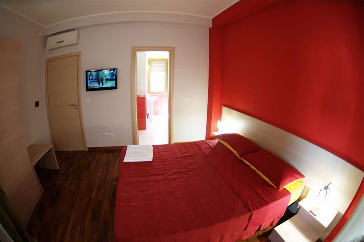 A bedroom with a red bedspread, a television on the wall, and a door leading to another room.