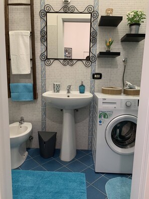 Et badeværelse med vask, bidet, vaskemaskine og hylder med toiletartikler.