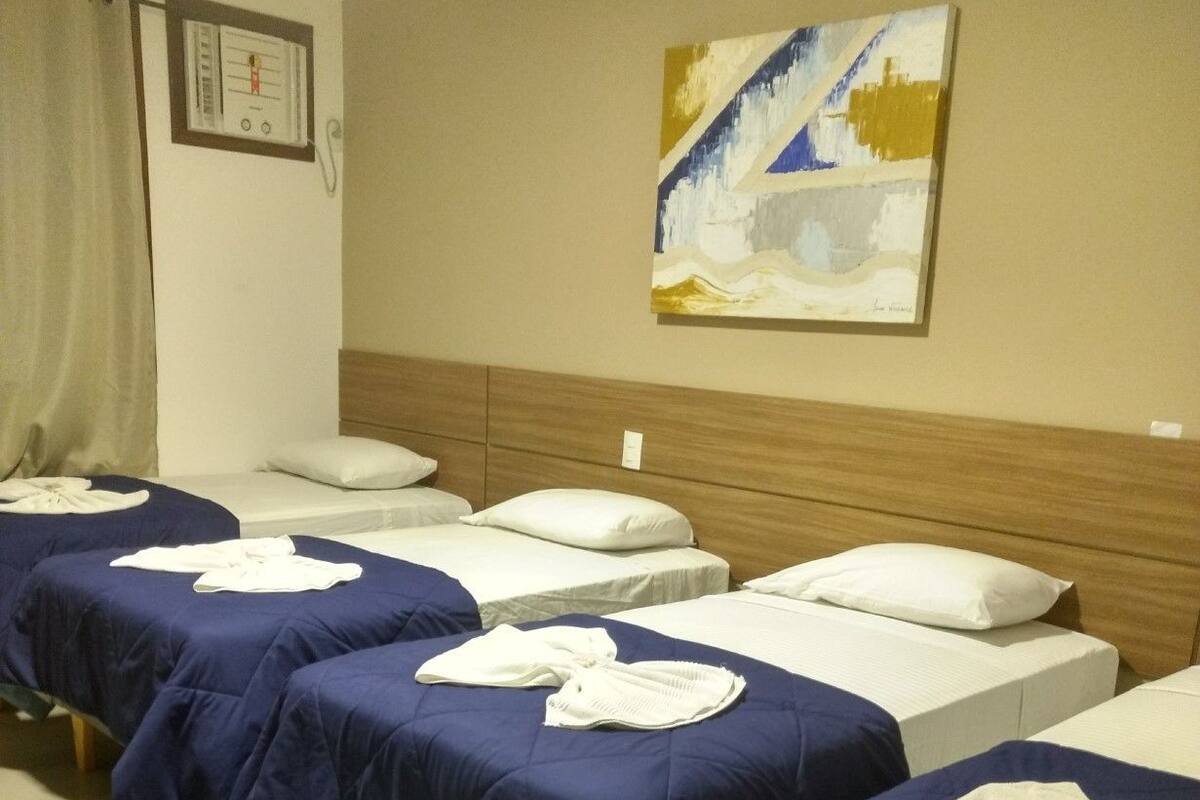 Quarto de hotel com três camas, cada uma com colcha azul e almofadas brancas. Há uma pintura na parede e um quadro emoldurado acima da porta.