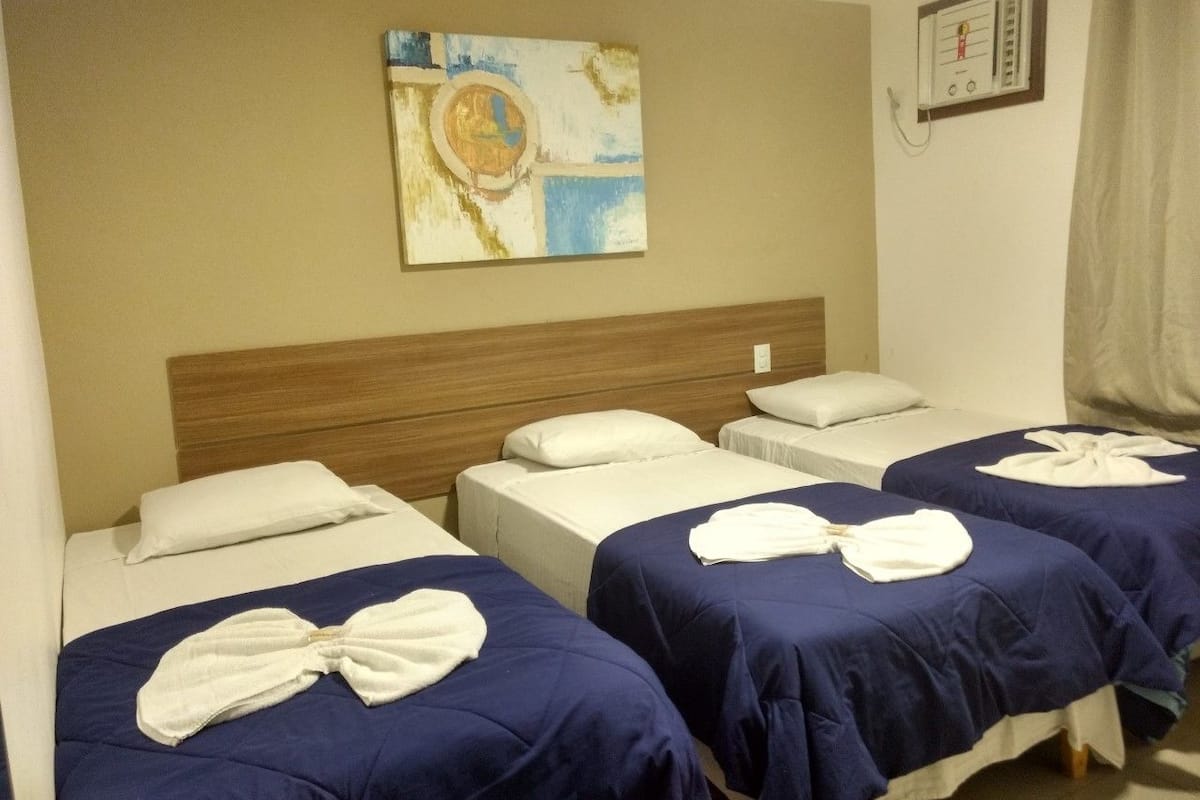 Quarto de hotel com três camas, cada uma com colcha azul e almofadas brancas. Há um quadro na parede acima das camas.