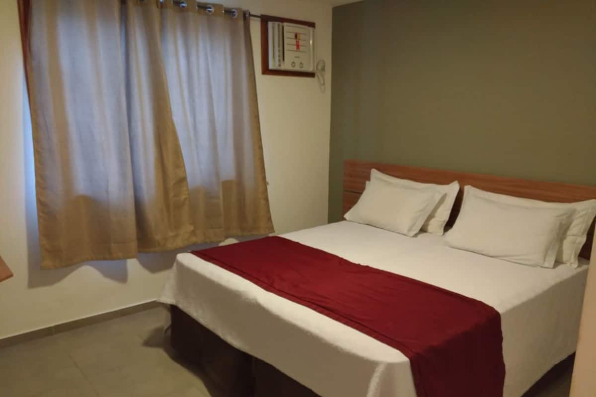 Uma cama de solteiro com colcha branca e vermelha, cabeceira de madeira e uma janela com cortinas bege.