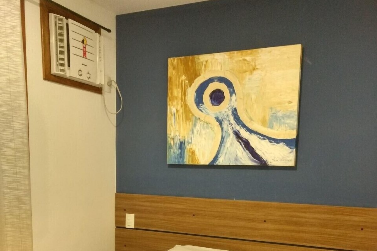 A hotel room with a bed, a painting on the wall, and a thermostat.