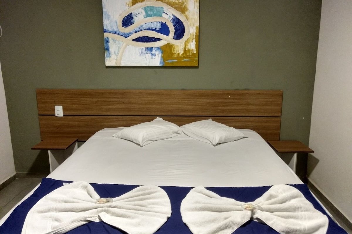Cama bem arrumada com cabeceira de madeira, manta azul e branca, e uma grande pintura abstrata na parede.