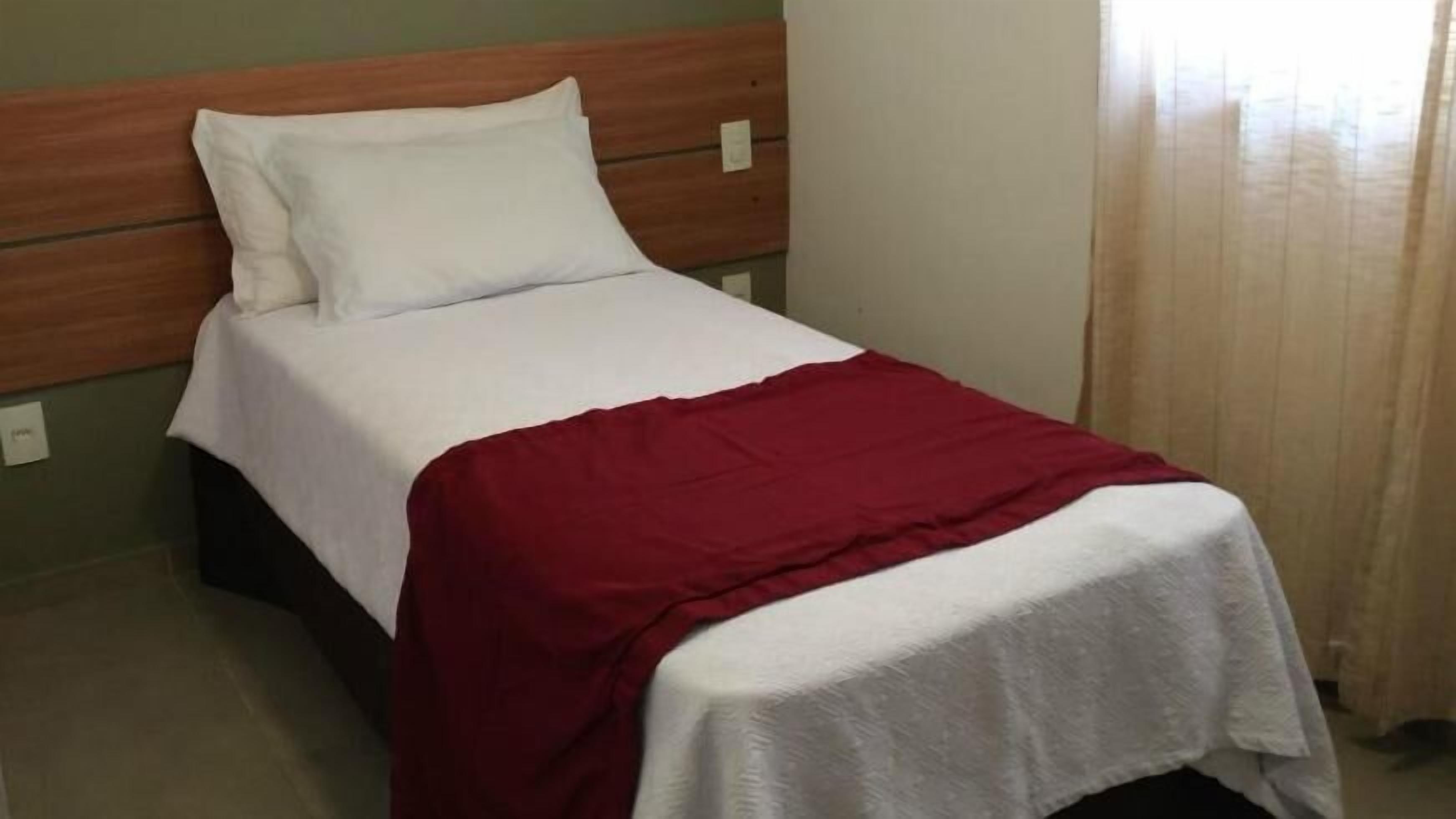 Una cama individual con cabecera de madera, ropa de cama blanca y una manta roja.