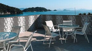 Een terras buiten met een betegelde mozaïek muur, witte stoelen, een glazen tafel en lichtjes, met op de achtergrond een helderblauwe lucht en een uitzicht op zee en heuvels in de verte.