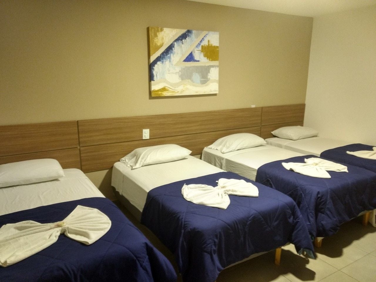 A hotel room with four single beds, each with a blue bedspread and white pillows. There is a painting on the wall above the beds.