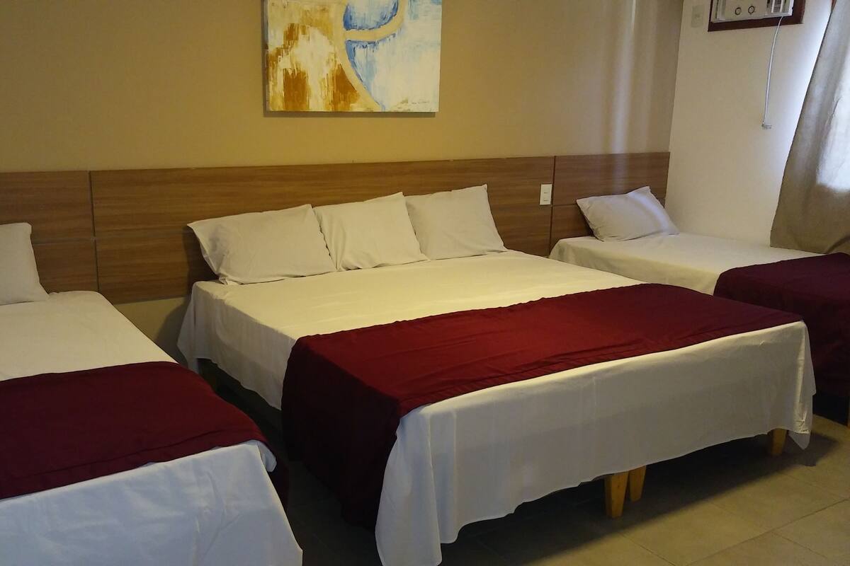Quarto de hotel com três camas, cada uma com roupa de cama branca e manta vermelha.