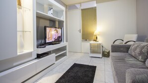 Uma sala de estar moderna com uma televisão de tela plana em uma unidade de entretenimento branca, um sofá cinza e uma pequena mesa lateral com uma luminária.