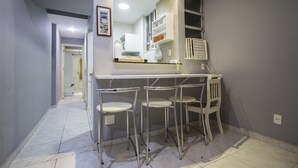 Uma cozinha compacta com bancada branca, quatro cadeiras de metal e forno embutido.