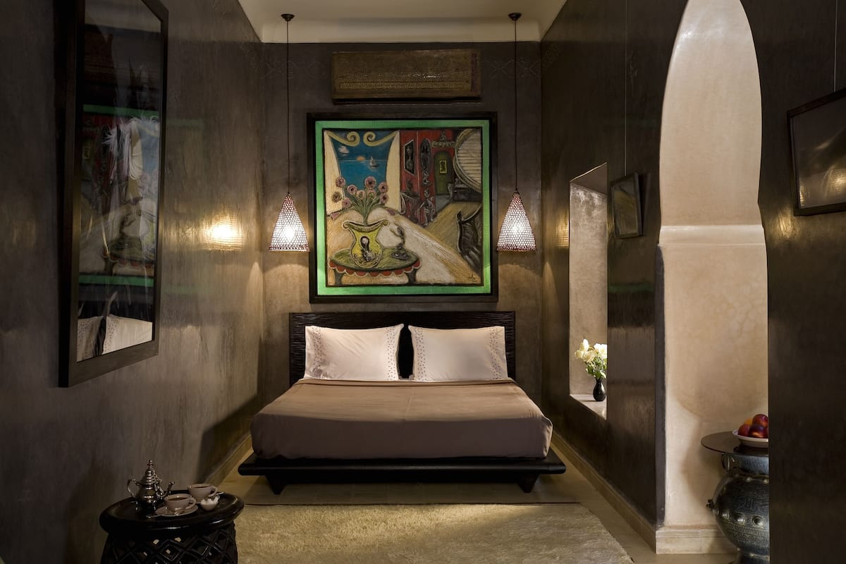 A bedroom with a large bed, a small table with a teapot and cups, a framed painting above the bed, and a vase with flowers on a shelf.