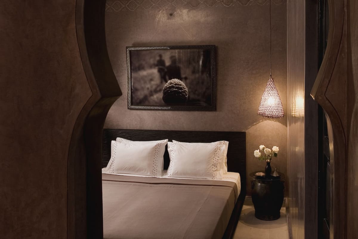 A bedroom with a bed, a framed picture on the wall, a small table with flowers, and a hanging light fixture.