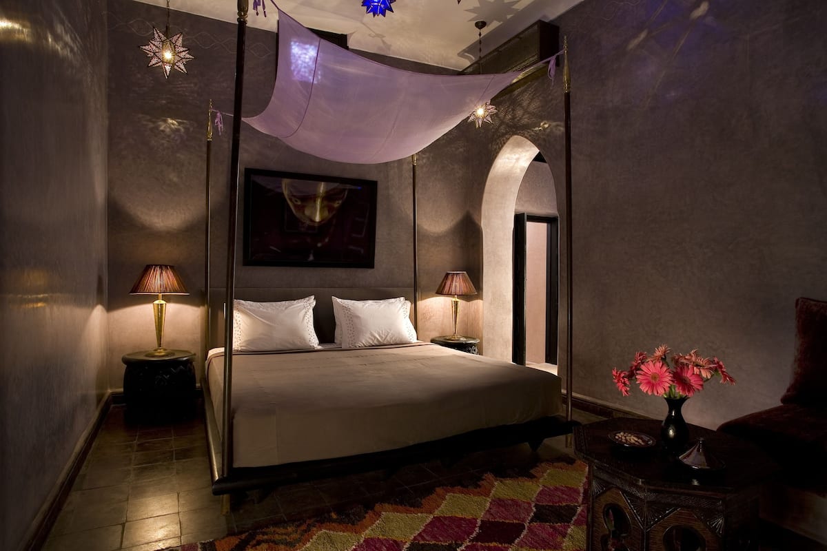 A four-poster bed with a canopy, two bedside lamps, a nightstand, a vase of flowers, and a framed artwork on the wall.