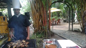 A chef grilling meat outdoors with a grill, a table with plates of vegetables, and a speaker in the background. - Hareesha Holiday Resort (Yakkalamulla)