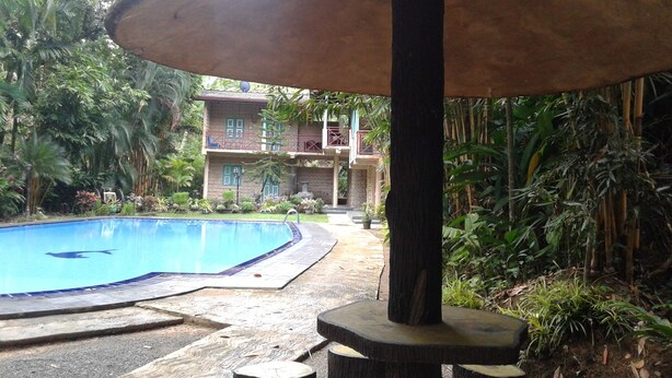 A pool area with a wooden structure overhead, surrounded by lush greenery and a two-story building in the background. - Hareesha Holiday Resort (Yakkalamulla)