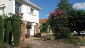 A white building with a red-tiled roof, a paved driveway, and various potted plants and shrubs.