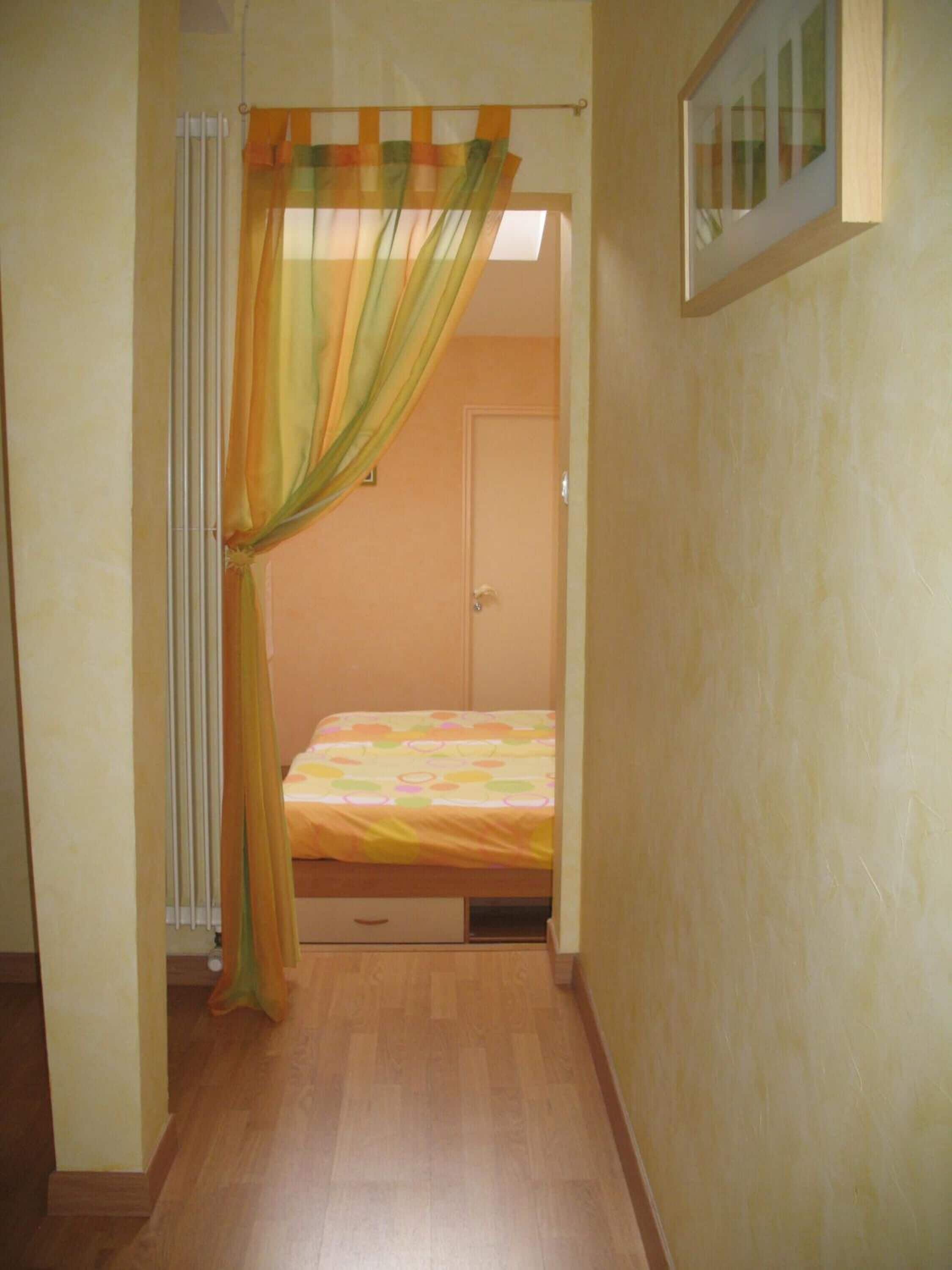 Un pasillo estrecho que da a un dormitorio con cama, un radiador y una puerta.