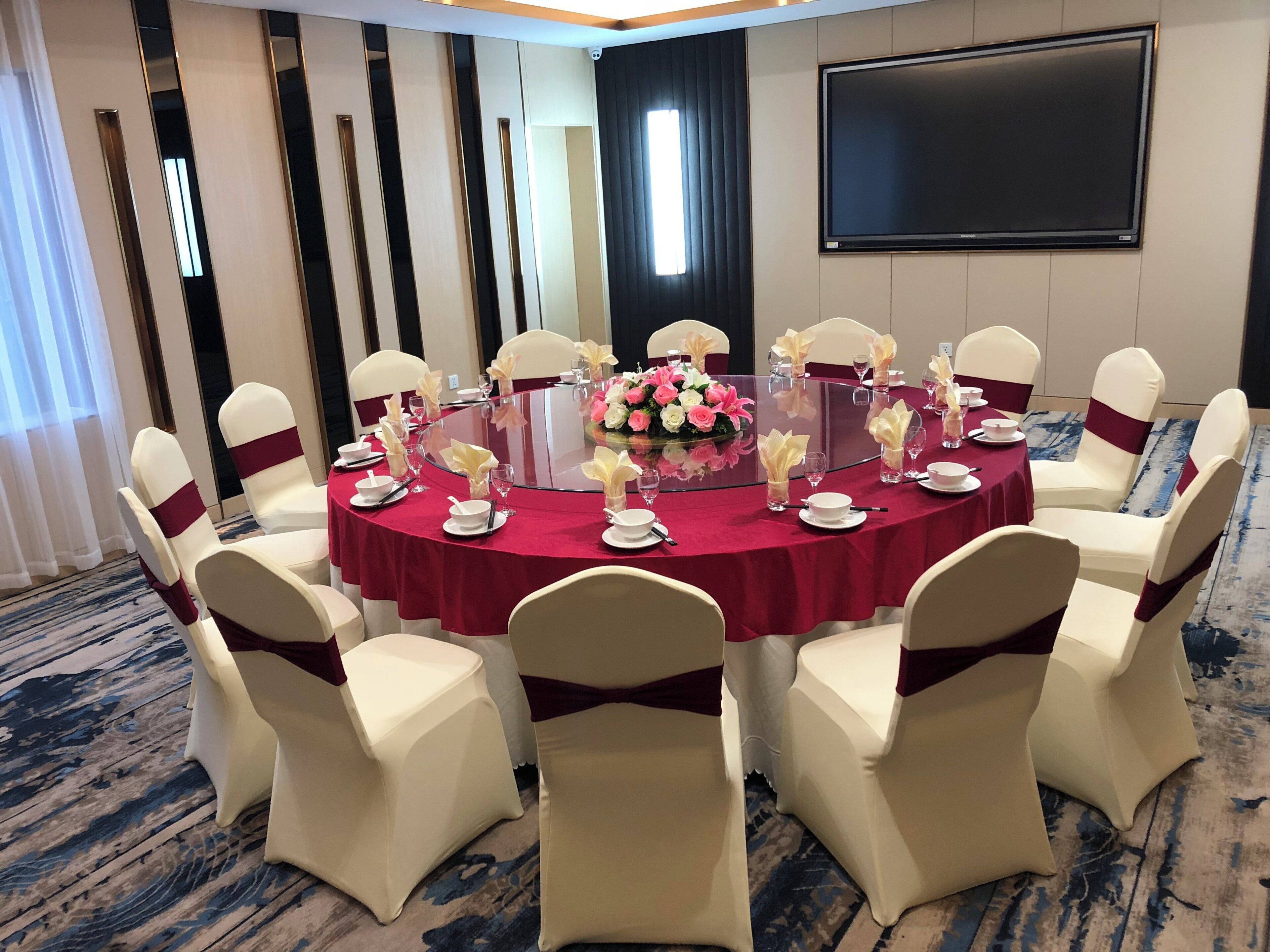 A conference room with a round table, white chairs with red sashes, and a large screen on the wall.