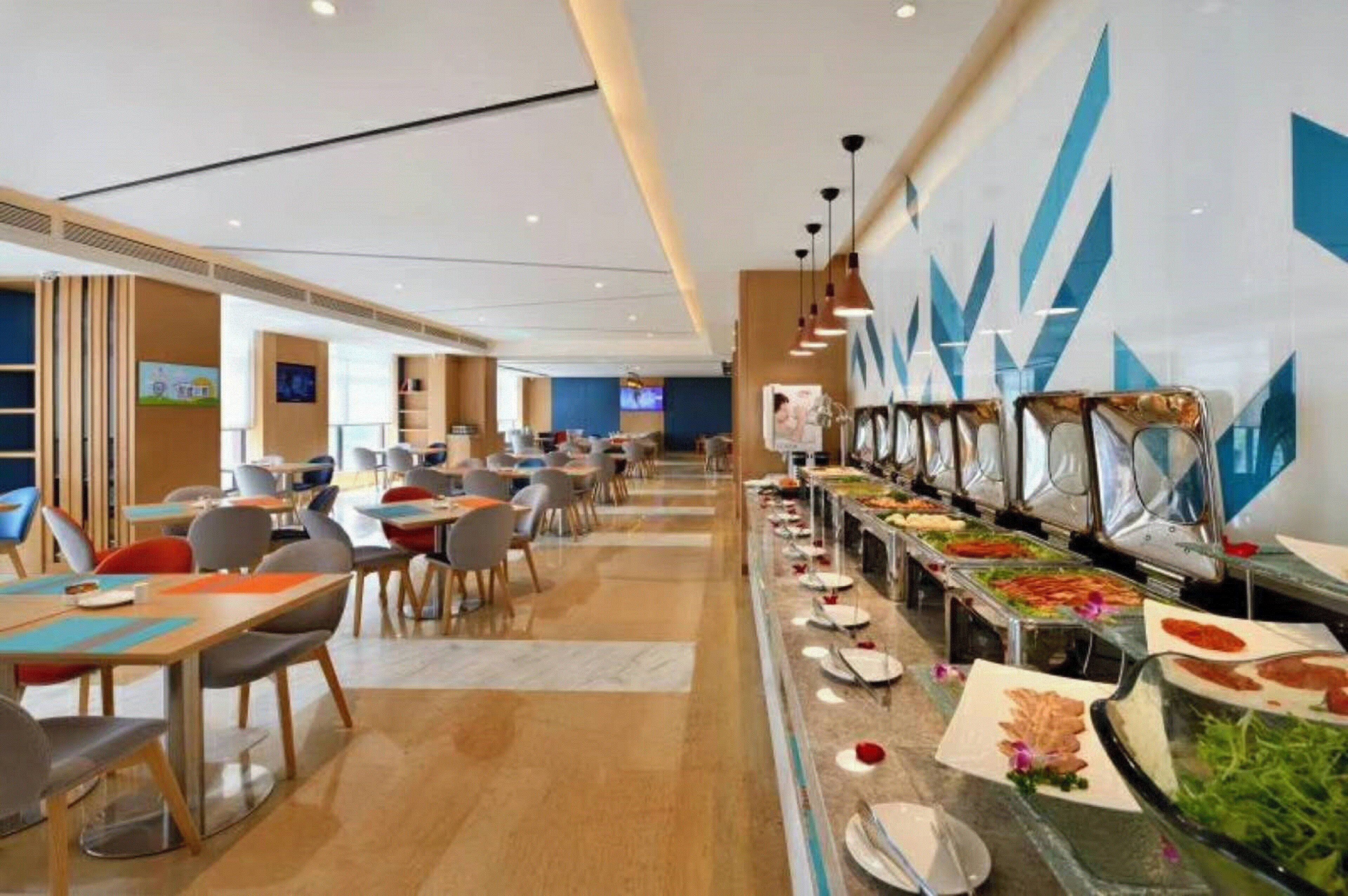 Un comedor moderno de hotel con varias mesas y sillas, una barra de buffet con diversas opciones de comida y una pared decorativa con motivos geométricos.
