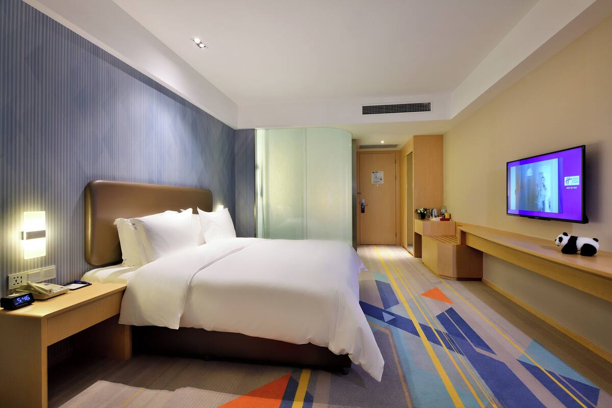 A hotel room with a large bed, a bedside table with a lamp, a TV on the wall, and a panda plush toy.