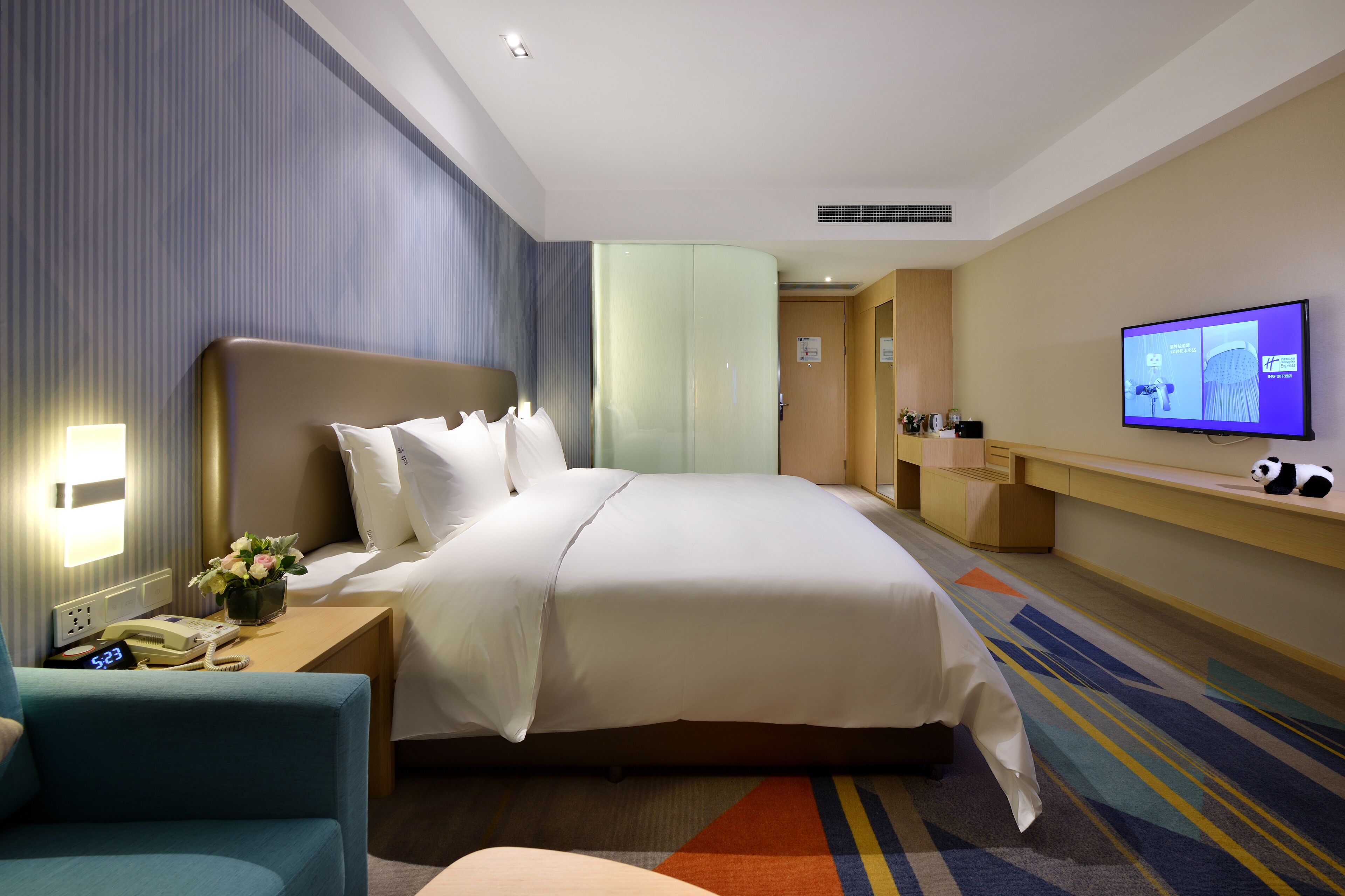 A hotel room with a large bed, a bedside table with a lamp, a TV, and a small desk with a chair.