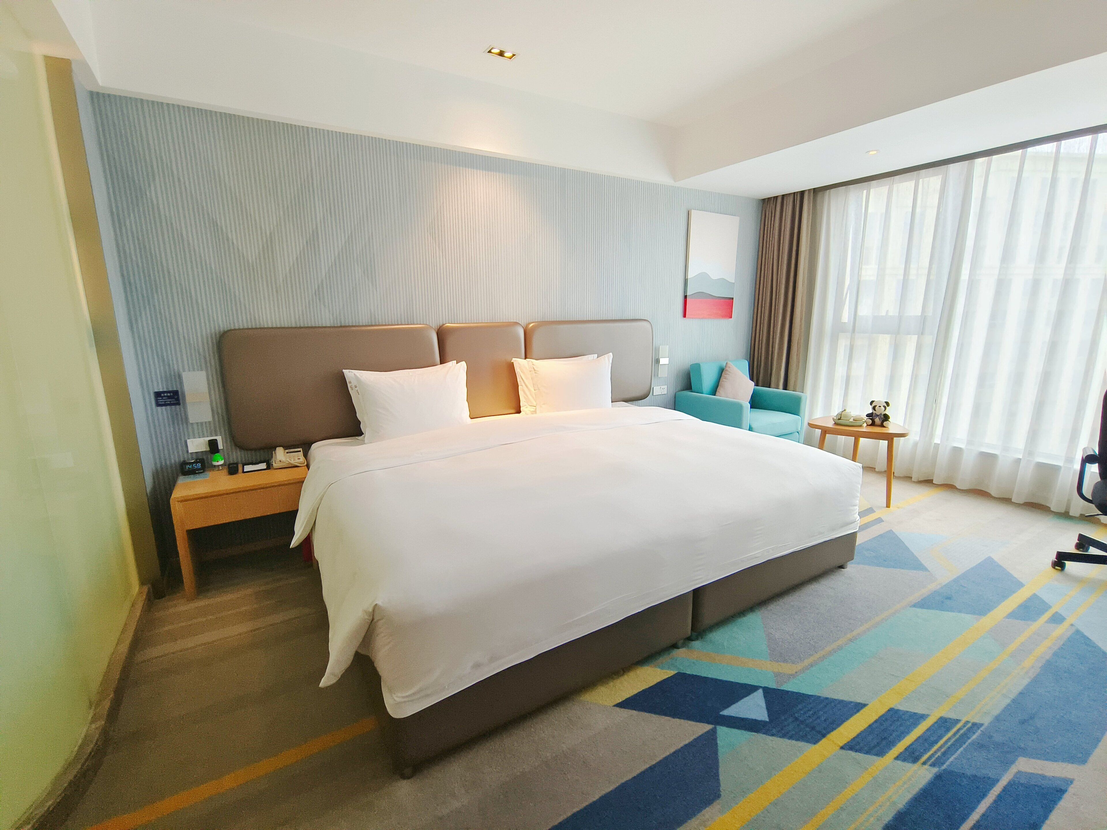 Habitación de hotel con una cama grande, una mesita de noche, una pequeña mesa, una silla y una ventana con cortinas.