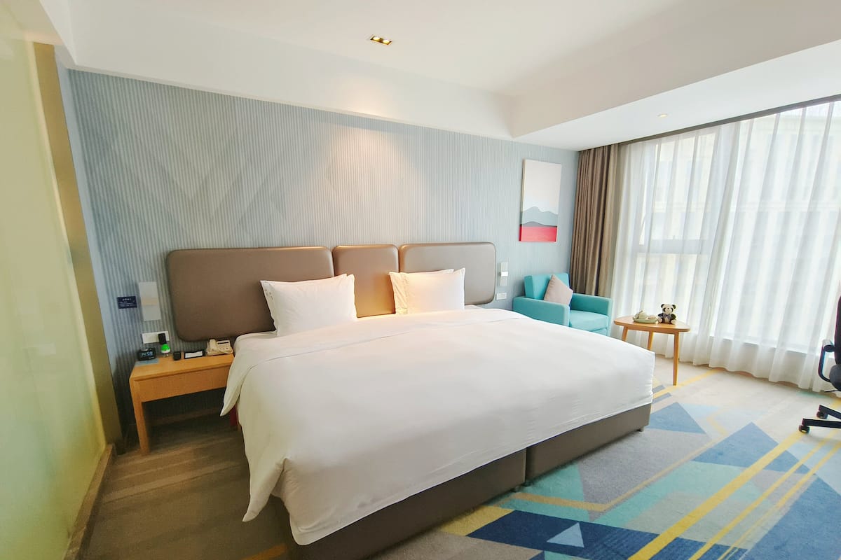 Une chambre d’hôtel avec un grand lit, une table de chevet, une petite table, une chaise et une fenêtre avec des rideaux.