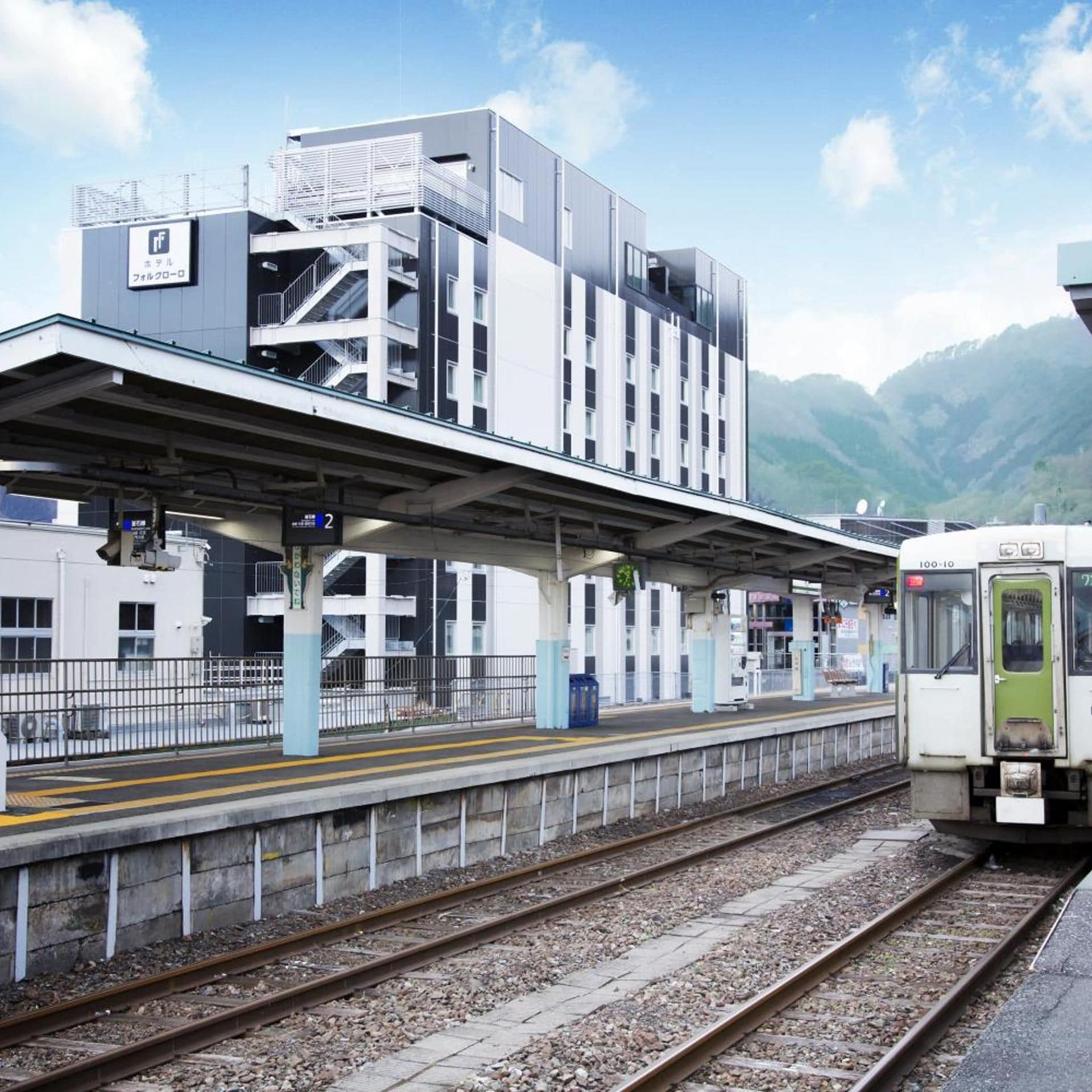 A train station platform with a modern building structure, a train on the tracks, and a mountainous backdrop.