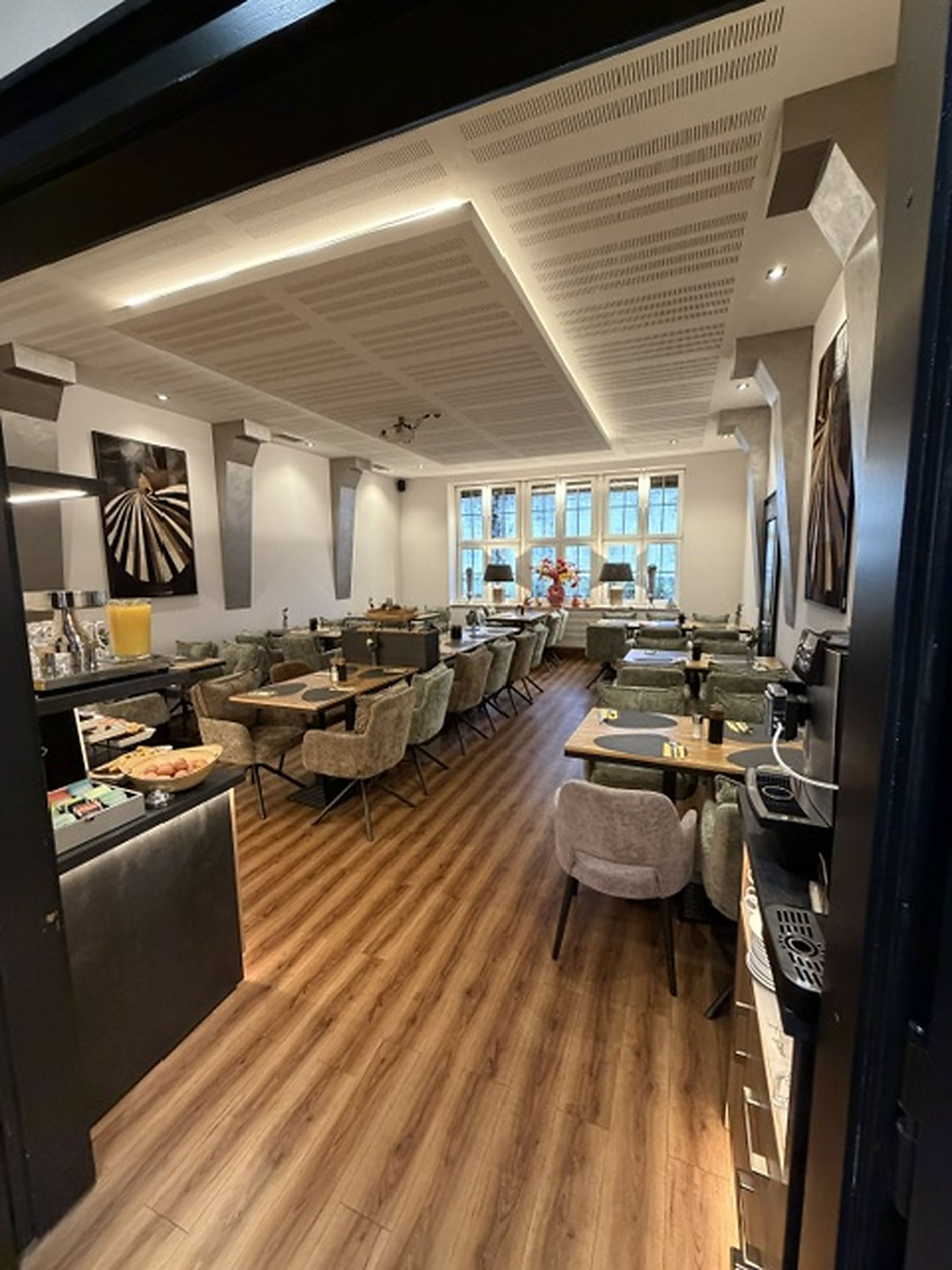 A modern dining area with wooden flooring, multiple seating arrangements, and large windows providing natural light.