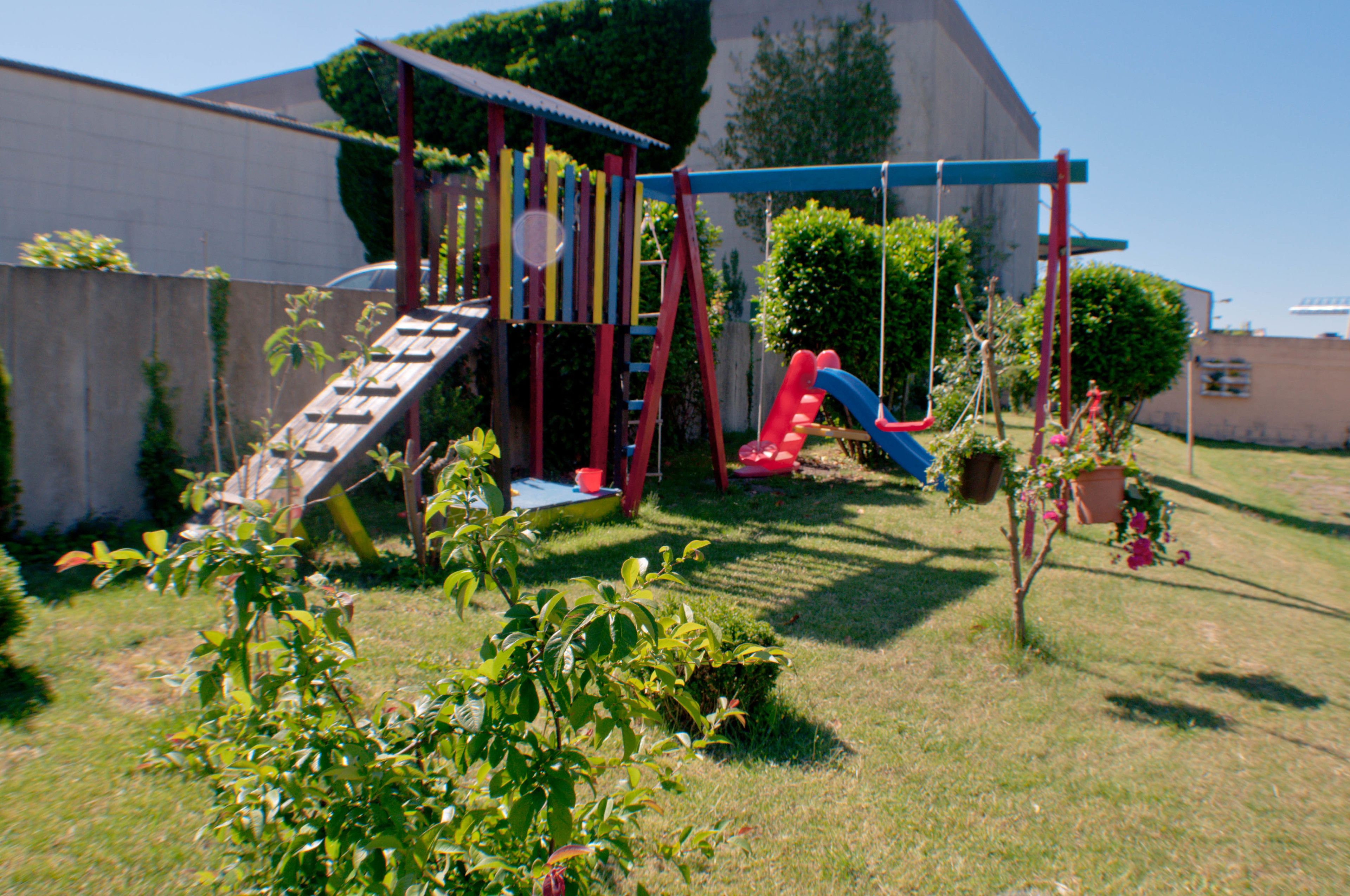 Un parque infantil con un tobogán y estructura para escalar, rodeado de césped y plantas en macetas.