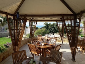An outdoor dining area with a wooden table and chairs, a pergola structure, and a view of the sea. - B&B La Rosa Dei Venti (Scicli)